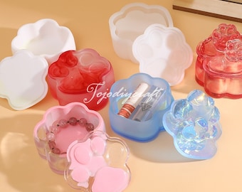 Può includere: Sei scatole di resina a forma di impronta di zampa con coperchi in vari colori, tra cui rosa, blu, bianco e rosso. Le scatole sono riempite con piccoli oggetti, come un braccialetto, rossetto e altri gingilli.