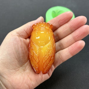 May include: A detailed amber-coloured cicada pendant with intricate carvings. The pendant is shaped like a cicada with wings and a head. The pendant is a beautiful example of insect jewellery.