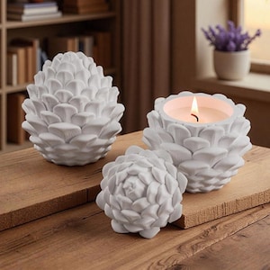 May include: Three white, decorative pine cone-shaped objects on a wooden surface. One is a candle holder with a lit candle, and the other two are solid sculptures. The objects are arranged in a group.