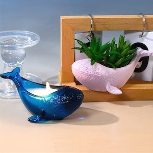 Puede incluir: Dos portavelas con forma de ballena. Uno es azul y contiene una vela blanca. El otro es rosa y contiene una pequeña planta verde suculenta.