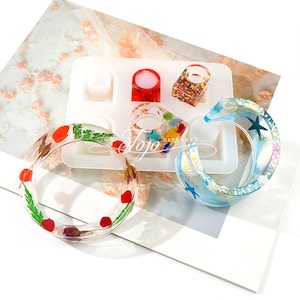 May include: Silicone mold for making resin bracelets and rings. The mold has two round bracelet shapes, one crescent moon shape, and several smaller ring shapes. The mold is shown with finished resin pieces, including a red and green floral bracelet, a blue and white seashell bracelet, and a red, green, and gold ring.