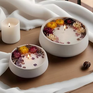 May include: Three lit candles. Two are white, bowl-shaped candles with dried flowers and petals. The third is a simple white pillar candle. The candles are on a wooden surface with white fabric in the background. The bowl candles have yellow, pink, and white flowers.