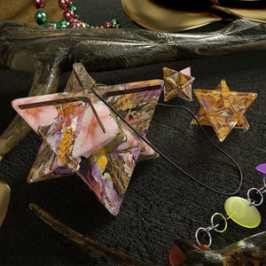 Op de afbeelding: Drie stervormige hangers met verschillende patronen. De grootste hanger is roze met een bloemenpatroon, de middelste hanger is bruin met een geometrisch patroon en de kleinste hanger is goud met een geometrisch patroon. De hangers hangen aan een zwart koord met zilveren kettingschakels en gekleurde kralen.
