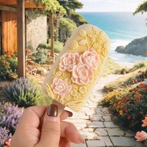 Może przedstawiać: Żółty lizak z kwiatowym wzorem. Lizak jest ozdobiony różowymi i żółtymi kwiatami i liśćmi. Lizak jest trzymany w dłoni na tle kamiennej ścieżki i plaży.