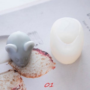 Op de afbeelding: Een witte siliconen mal in de vorm van een muis. De mal ligt op een wit oppervlak naast een grijs muisvormig object.