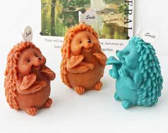May include: Three hedgehog-shaped candles, one brown, one teal blue, and one light brown, each holding a mushroom. The candles have a small flag on top that says "Smile".