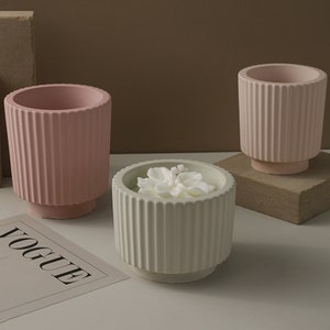 Puede incluir: Tres portavelas decorativos con diseño acanalado. Dos son rosas y uno es blanco con una vela de flor blanca en su interior.