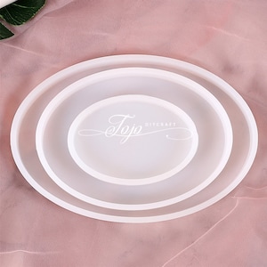 May include: Three white oval silicone moulds for resin casting. The moulds are nested inside each other, with the largest mould on the bottom and the smallest mould on top. The moulds are on a pink fabric background. The text "Jojo DIYCRAFT" is printed on the largest mould.