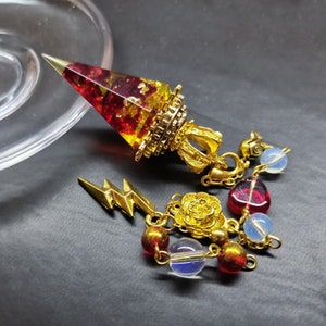 May include: A gold and red crystal pendulum with a chain and charms. The charms include a lightning bolt, a rose, and red and clear glass beads.