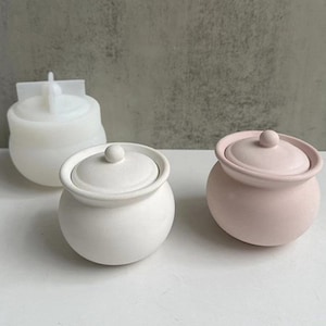 Puede incluir: Tres tarros de cerámica con tapa, uno blanco, uno rosa y uno blanco con una tapa ligeramente descentrada. Los tarros son redondos y tienen una forma ligeramente bulbosa.