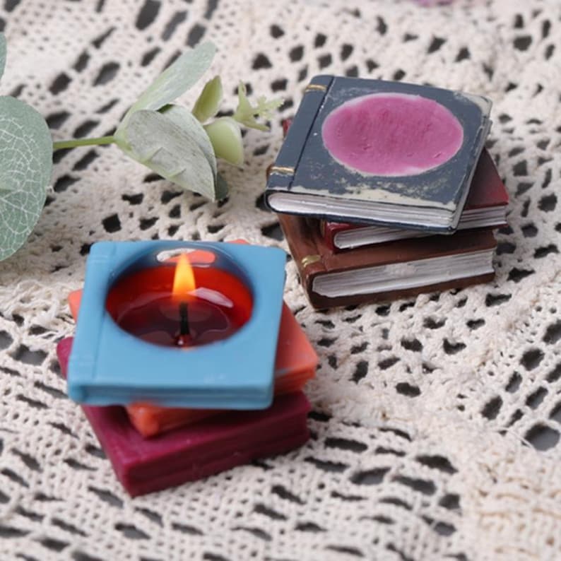 May include: A set of three stacked book-shaped candles. The top candle is blue with a red wax pool and a lit wick. The bottom two candles are red and orange.