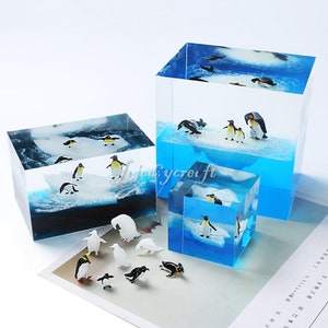 Puede incluir: Tres cajas de acrílico transparente con pingüinos e icebergs en su interior. Las cajas son de diferentes tamaños y formas. También hay varias pequeñas figuras de pingüinos esparcidas sobre una superficie blanca.