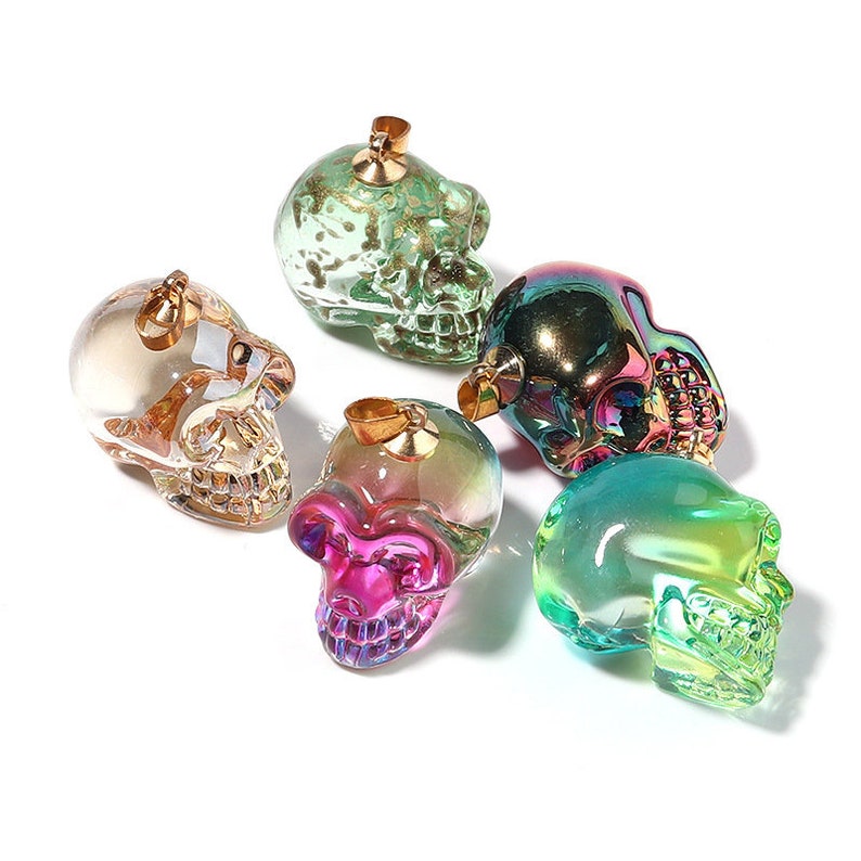 K&ouml;nnte beinhalten: Sechs regenbogenfarbene Glassch&auml;del-Anh&auml;nger mit goldfarbenen Metallscharnieren. Die Sch&auml;del sind in verschiedenen Farben, darunter Gr&uuml;n, Blau, Lila und Pink.
