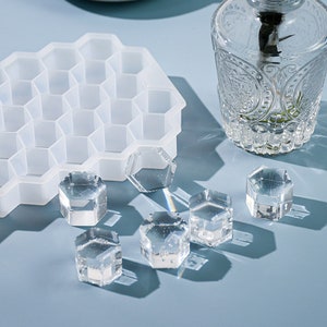 Può includere: Uno stampo in silicone a forma di favo con sei cavità esagonali. Sei pezzi esagonali in resina trasparente sono sparsi su una superficie blu chiaro.