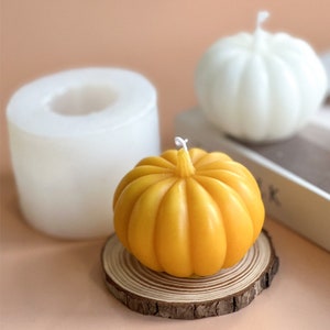 May include: A yellow pumpkin-shaped candle with a white wick sits on a wooden slice. A white pumpkin-shaped candle is in the background.