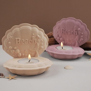 May include: Two decorative candle holders shaped like seashells with the word "Beach" embossed on them. The candle holders are in pink and beige and have a mermaid tail design.