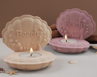 Op de afbeelding: Twee decoratieve kaarshouders in de vorm van schelpen met het woord "Beach" erop gegraveerd. De kaarshouders zijn roze en beige en hebben een zeemeerminstaartontwerp.
