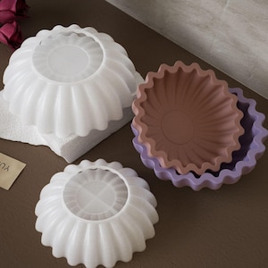 May include: Three white silicone cake moulds with fluted edges. The moulds are in different sizes and are arranged on a light grey surface.
