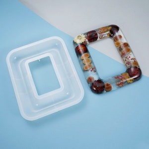 May include: A clear silicone mold for making resin picture frames. The mold is rectangular with a smaller rectangular opening in the center. A finished resin picture frame with dried flowers and berries is shown next to the mold.