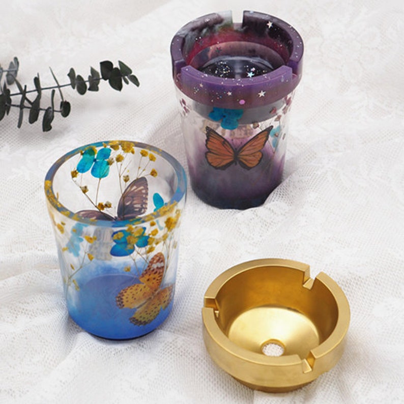 May include: Three different ashtrays. One is a clear glass ashtray with a blue and yellow butterfly design and dried flowers. Another is a clear glass ashtray with a purple and pink galaxy design. The third is a gold metal ashtray with a round top and a small hole in the center.