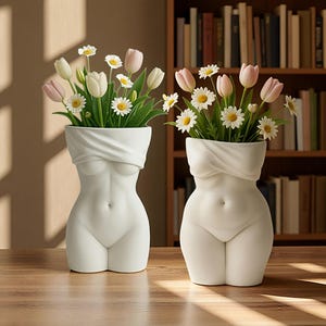 May include: Two white ceramic vases shaped like human torsos, each featuring a draped fabric design across the chest. Both vases are filled with a bouquet of white daisies and light pink tulips, creating a floral arrangement in each vessel.