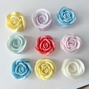 May include: Nine handmade rose-shaped candles in various pastel colors, including light blue, yellow, pink, and lavender. The candles are arranged in a circle on a white surface.