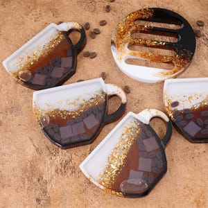 May include: Set of four coffee cup-shaped coasters with a white, brown, and gold resin design. The coasters feature a coffee bean and gold flake design.