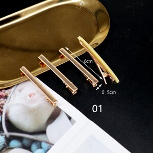 K&ouml;nnte beinhalten: Vier goldene Haarklammern, jede 15,2 cm lang und 1,3 cm breit. Die Klammern sind auf einem goldenen Tablett mit wei&szlig;em Hintergrund angeordnet.