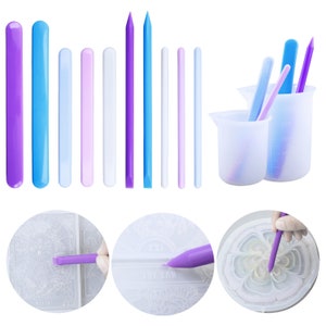 May include: A set of 10 silicone tools for resin art, including a variety of shapes and sizes. The tools are purple, blue, and white. The set also includes two silicone measuring cups.