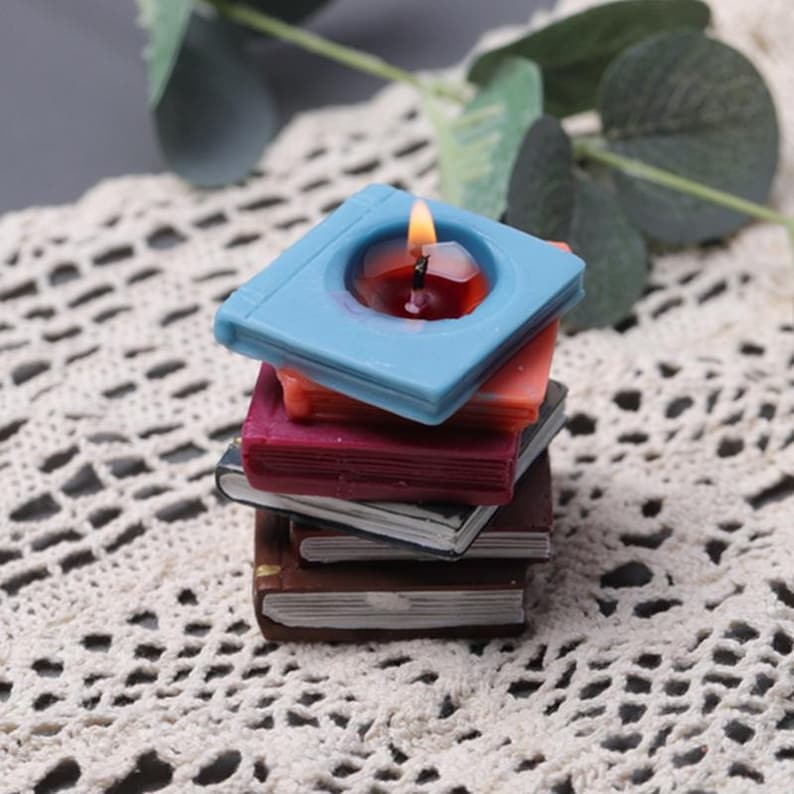 May include: A stack of book-shaped candles, the top one is lit and blue with a red wax pool. The candles are stacked on a white crocheted surface.
