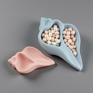 May include: A light blue seashell-shaped dish filled with small white and pale pink pearls, accompanied by an empty matching light pink seashell-shaped dish. Both dishes feature a smooth matte finish and a divided interior structure.