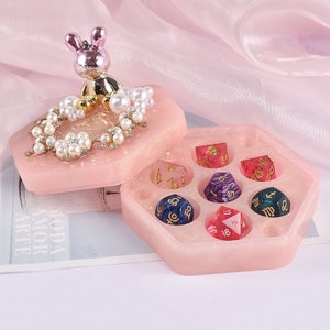May include: A pink hexagonal dice box with a decorative lid featuring a bunny figurine, pearls, and floral accents. The box contains seven colorful polyhedral dice with gold numbering and zodiac symbols.
