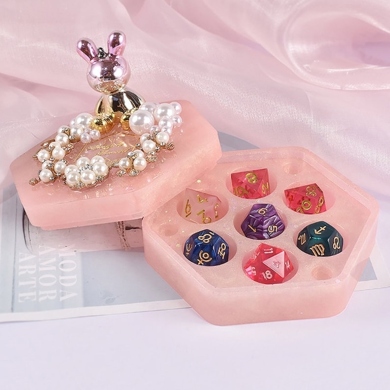 May include: A pink hexagonal dice box with a decorative lid featuring a bunny figurine, pearls, and floral accents. The box contains seven colorful polyhedral dice with gold numbering and zodiac symbols.