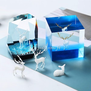 May include: Two clear resin paperweights with a deer figurine inside. The paperweights are on a light blue surface. One paperweight is a cube shape and the other is a geometric shape.