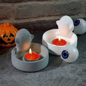 Könnte beinhalten: Zwei Halloween-Kerzenhalter, einer grau und einer weiß, jeweils mit einer Geisterfigur und einer kleinen roten Kerze. Der weiße Halter hat außerdem eine weiße Augenkugel-Figur.