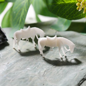 May include: Two white plastic miniature horses with saddles. The horses are standing on a gray surface.