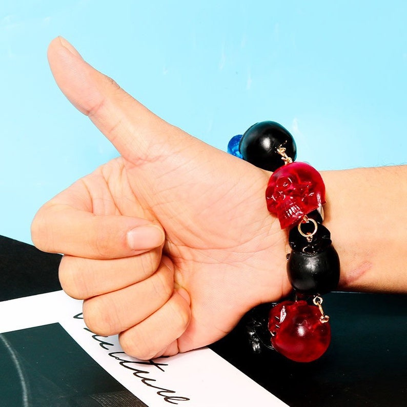 K&ouml;nnte beinhalten: Ein Armband mit schwarzen und roten Totenkopfperlen und goldenen Kettengliedern. Das Armband wird am Handgelenk einer Person getragen.