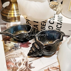 May include: Two black seashell-shaped ceramic dishes with gold glitter accents. The dishes are on a brown surface with a book and other decorative items.