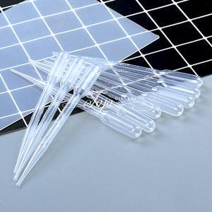 May include: A set of ten clear plastic droppers with a pointed tip and a bulb on the top. The droppers are arranged in a pile on a white and black checkered surface.