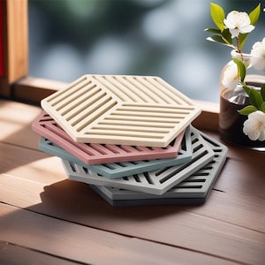 May include: A stack of six hexagonal silicone coasters in various colors, including beige, pink, blue, and gray. The coasters have a geometric pattern of raised lines.