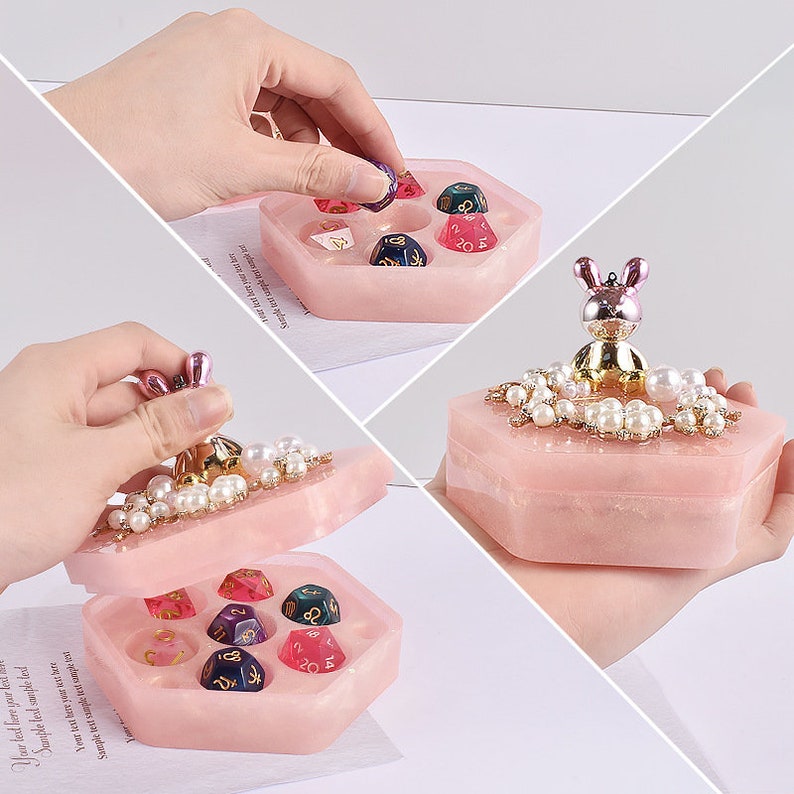 May include: A pink hexagonal dice box with a bunny figurine on top, adorned with pearls. The box is open, revealing several colorful dice. The box is made of a translucent material.