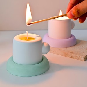 May include: Two ceramic cup-shaped candles, one white and one light purple, sit on matching colored saucers. A lit match is held over the purple candle. The candles have a yellow wax interior and a small handle. The saucer is a circular shape.