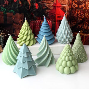 Puede incluir: Surtido de ocho velas con forma de árbol de Navidad en varios colores y diseños. Las velas están hechas de cera y tienen un acabado liso y pulido. Las velas están dispuestas en fila sobre una superficie blanca.