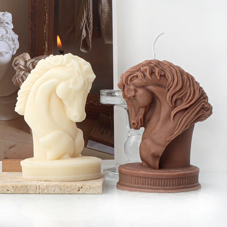 May include: Two decorative horse head candles, one white and one brown, with a classic design. The candles are on a light brown surface.