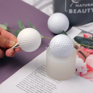 Puede incluir: Tres moldes de silicona en forma de bola de golf blanca. Los moldes están sobre una superficie blanca con un libro en el fondo. El libro tiene texto que dice "Donde los bosques verdes están aquí, aquí está el pequeño oriole amarillo que puede cantar."