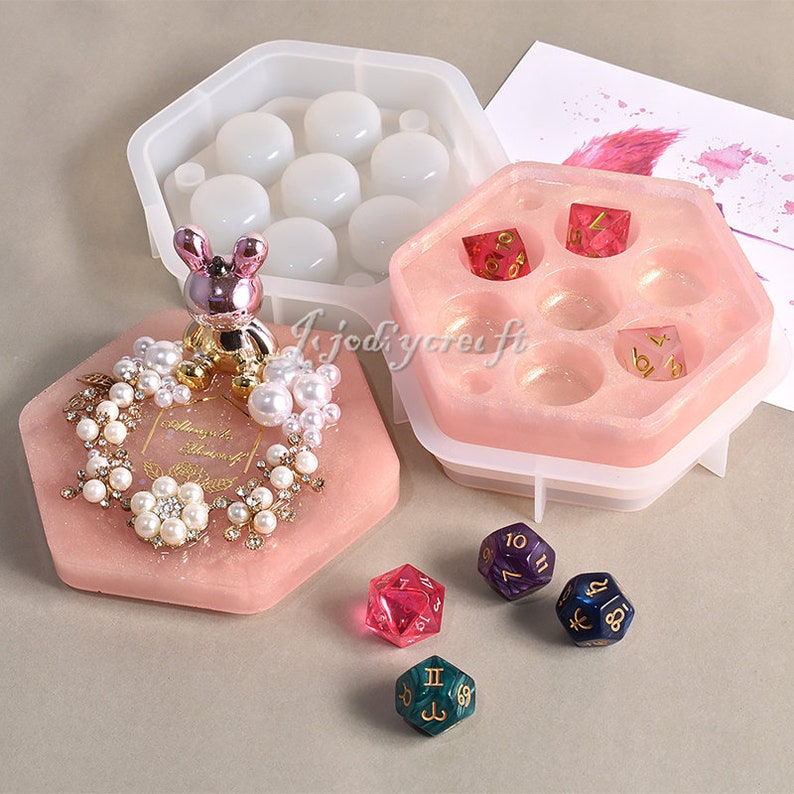May include: A collection of resin molds and dice. The molds are in various shapes, including a hexagonal tray with dice, a round mold, and a hexagonal tray with a decorative bunny and pearl embellishments. Several colorful dice are also present.