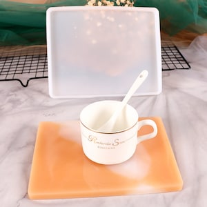 May include: A white ceramic mug with gold trim and the text "Recherch&eacute; Snow" and "Bonechina" on the side. The mug is sitting on a light orange and white resin coaster.