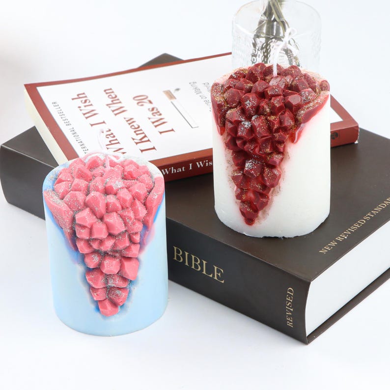 May include: Two decorative pillar candles with a crystal-like design. The candles are white with a red crystal-like design on the side. The candles are sitting on a black book with the word "BIBLE" printed on the cover.