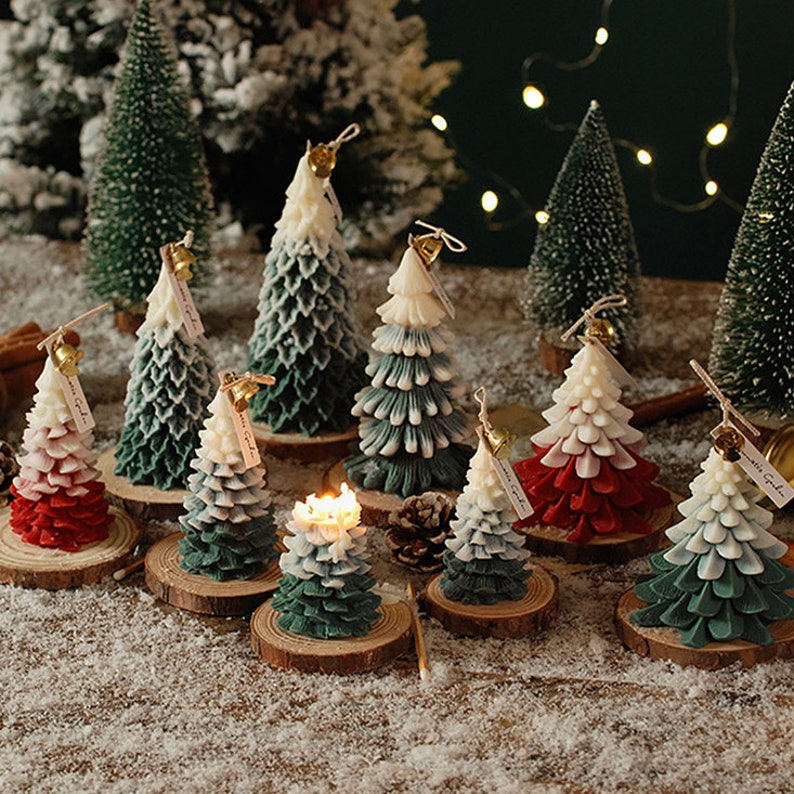 May include: Assortment of green, white, and red Christmas tree candles with gold bells and wooden bases. The candles are arranged on a snowy surface.