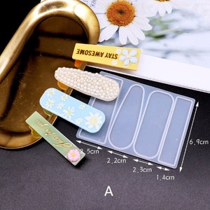 K&ouml;nnte beinhalten: Eine transparente Silikonform mit vier rechteckigen Vertiefungen zum Herstellen von Harzklammern f&uuml;r Haare. Die Form misst 6,9 cm x 2,5 cm. Die Form wird mit vier fertigen Harzklammern gezeigt, jede mit einem anderen Design. Eine Klammer hat ein wei&szlig;es Perlmuster, eine hat ein blaues und wei&szlig;es G&auml;nsebl&uuml;mchenmuster, eine hat ein gr&uuml;nes und rosa G&auml;nsebl&uuml;mchenmuster und eine hat einen gelben Hintergrund mit dem Text "Stay Awesome" und einem wei&szlig;en G&auml;nsebl&uuml;mchen.
